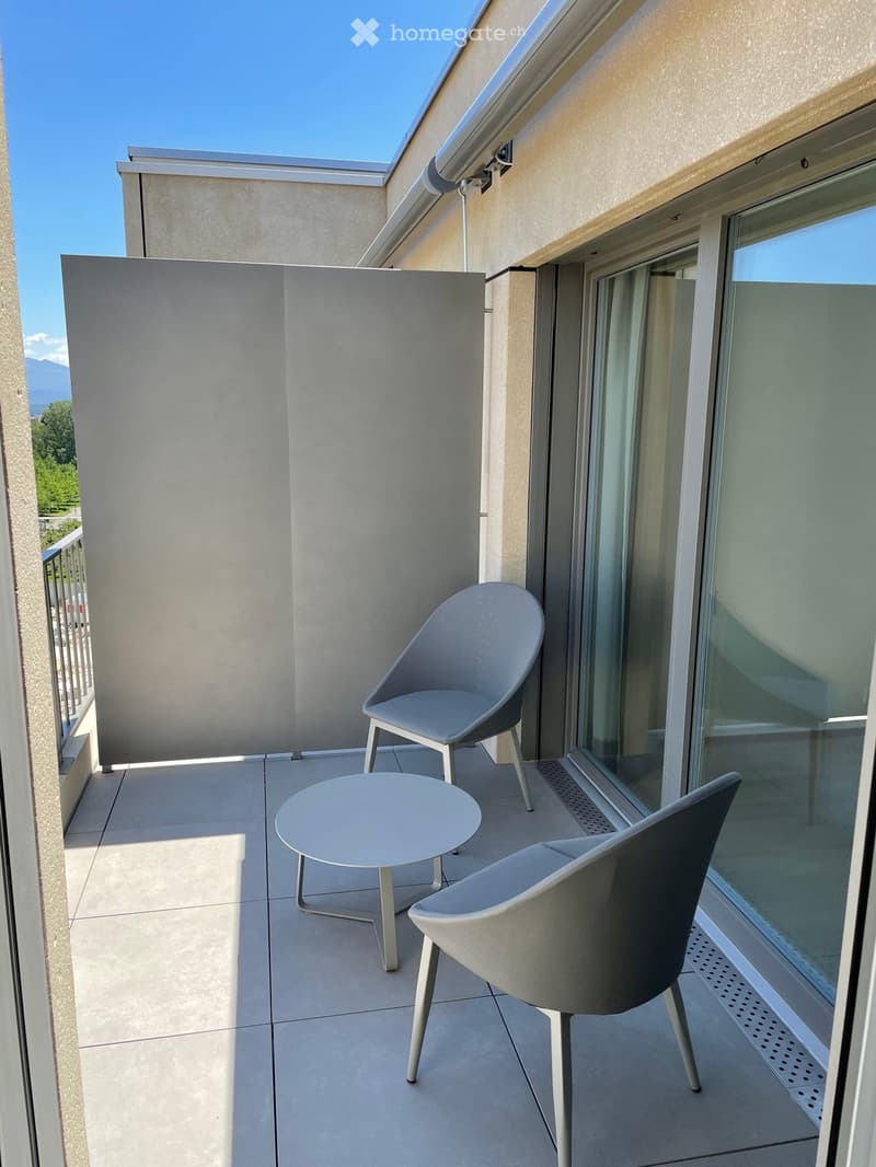 Modern balcony with tiled floor, two contemporary chairs, and a round table, overlooking a clear blue sky and distant mountains.