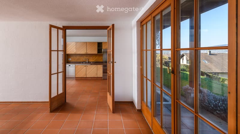 Open-plan space with terracotta tiled floor, wooden kitchen cabinets, sink, dishwasher, and oven visible through an open doorway. Large wooden-framed glass doors lead to a view of a garden and neighboring houses.