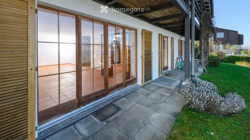 Outdoor patio area with large glass doors leading to a tiled terrace, wooden shutters, and a well-maintained lawn with shrubs.