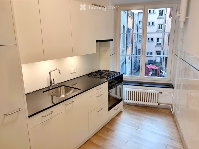 Modern kitchen with white cabinets, black countertop, stainless steel sink, gas stove, and oven. Wooden flooring and large window with view of the street.