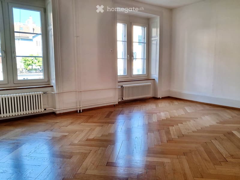 Empty room with wooden flooring, white walls, and large windows with radiators beneath them.