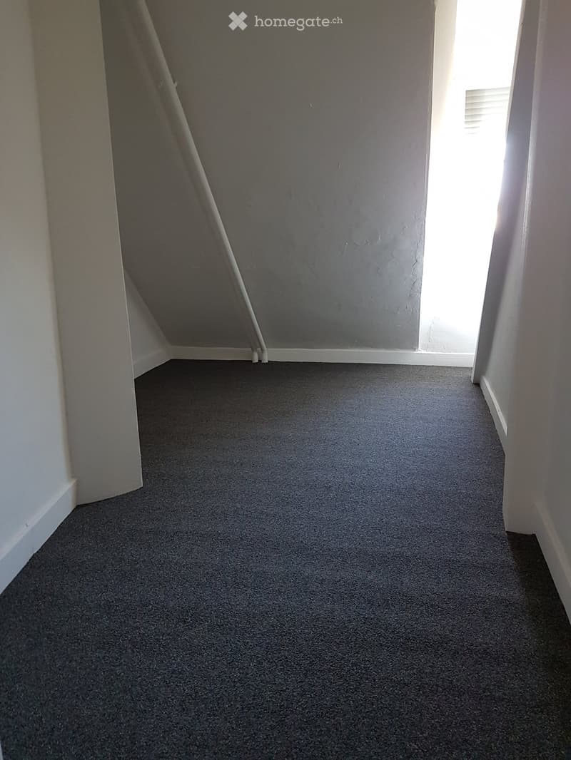 Empty hallway with grey carpet and white walls, featuring a window at the end allowing natural light to enter.
