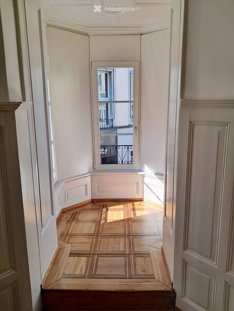 A narrow hallway with wooden flooring and white walls, featuring a window at the end that offers a view of a balcony with metal railings.