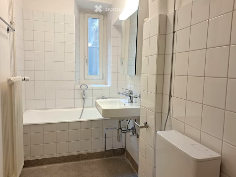 Modern bathroom with white tiled walls and floor, featuring a bathtub, sink, mirror, and toilet.