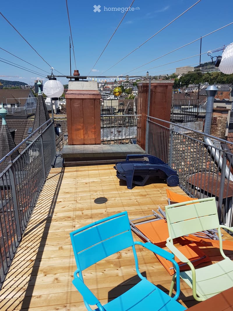 Wooden deck with metal railing, featuring colorful chairs and a table, overlooking a cityscape with visible rooftops and chimneys.