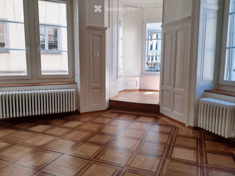 Empty room with wooden parquet flooring, white walls, radiators, and large windows offering a view of a balcony area.