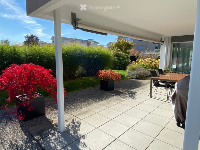 Patio area with tiled flooring, a wooden table, and chairs, surrounded by greenery and potted plants.