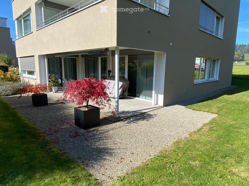 Modern house exterior with a covered patio area, large windows, and a well-maintained garden with potted plants and a gravel pathway.