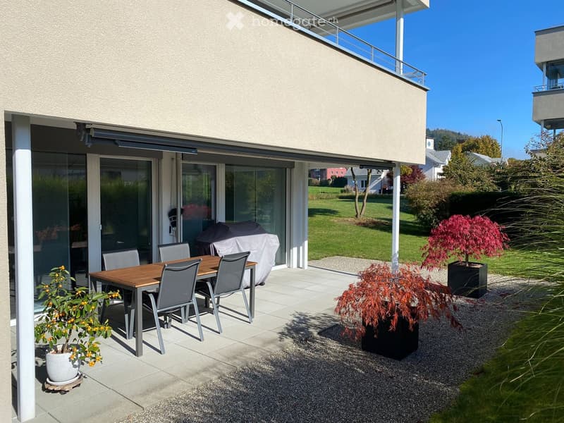Outdoor patio with a wooden dining table, four chairs, and a covered umbrella, surrounded by potted plants and a well-maintained lawn.