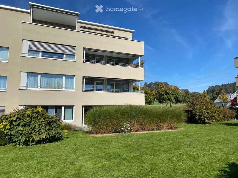 Modern two-story house with beige facade, large windows, and a balcony on the upper floor. The house is surrounded by a well-maintained lawn and shrubs.