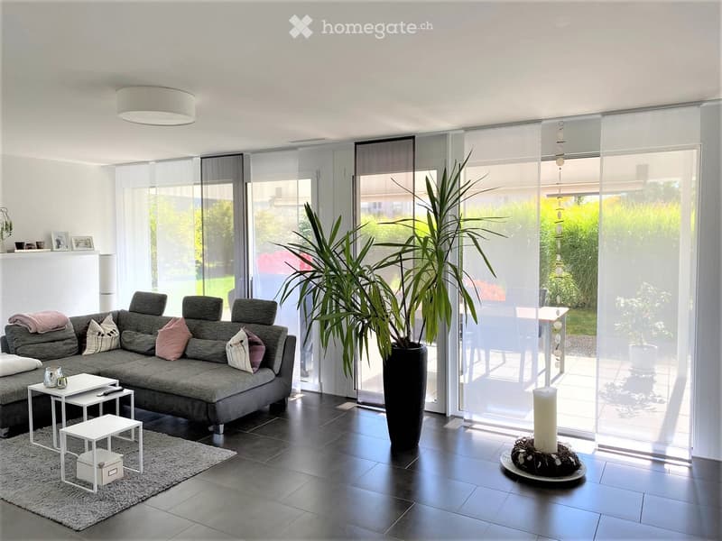 Modern living room with grey sectional sofa, white coffee table, small white stools, large potted plant, and a view of the garden through large glass doors.