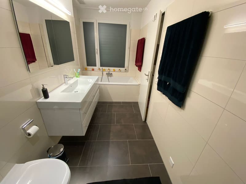 Modern bathroom with double sink, bathtub, toilet, large mirror, and towels hanging on the walls.
