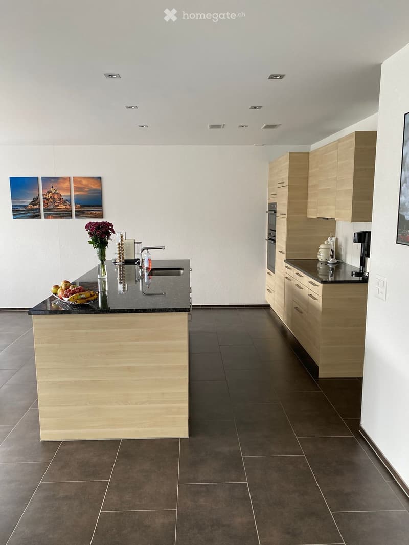 Modern kitchen with light wooden cabinets, dark countertops, and a central island. Features include a sink with a modern faucet, a coffee maker, a vase with flowers, and a fruit bowl on the island.