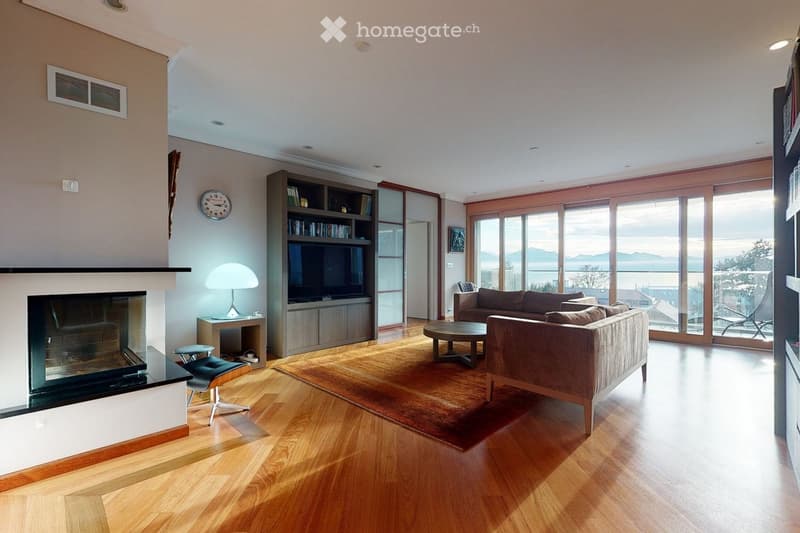 Modern living room with hardwood floors, large windows offering a scenic view, a fireplace, a sofa, a coffee table, a lamp, a bookshelf, and a small table with a stool.