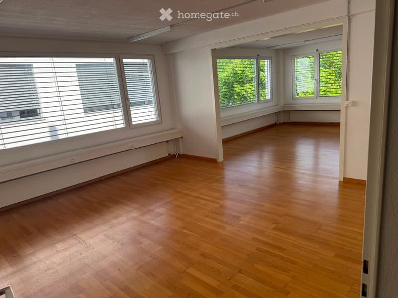 Empty room with wooden flooring, large windows with blinds, and white walls.