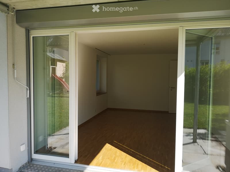 Empty room with wooden flooring, large glass sliding doors leading to an outdoor area, and visible sunlight streaming in.