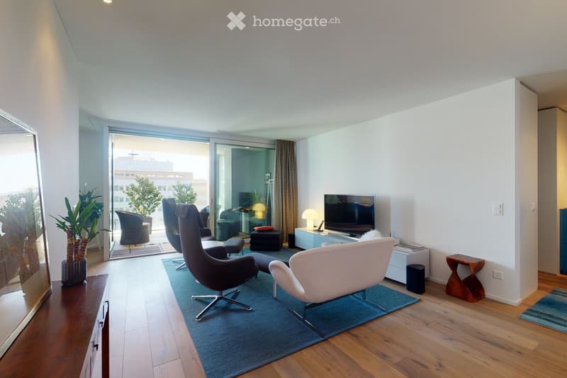 Modern living room with wooden flooring, large sliding glass doors leading to a balcony, a blue rug, a white armchair, a black leather chair, a small wooden side table, a television, and a lamp.