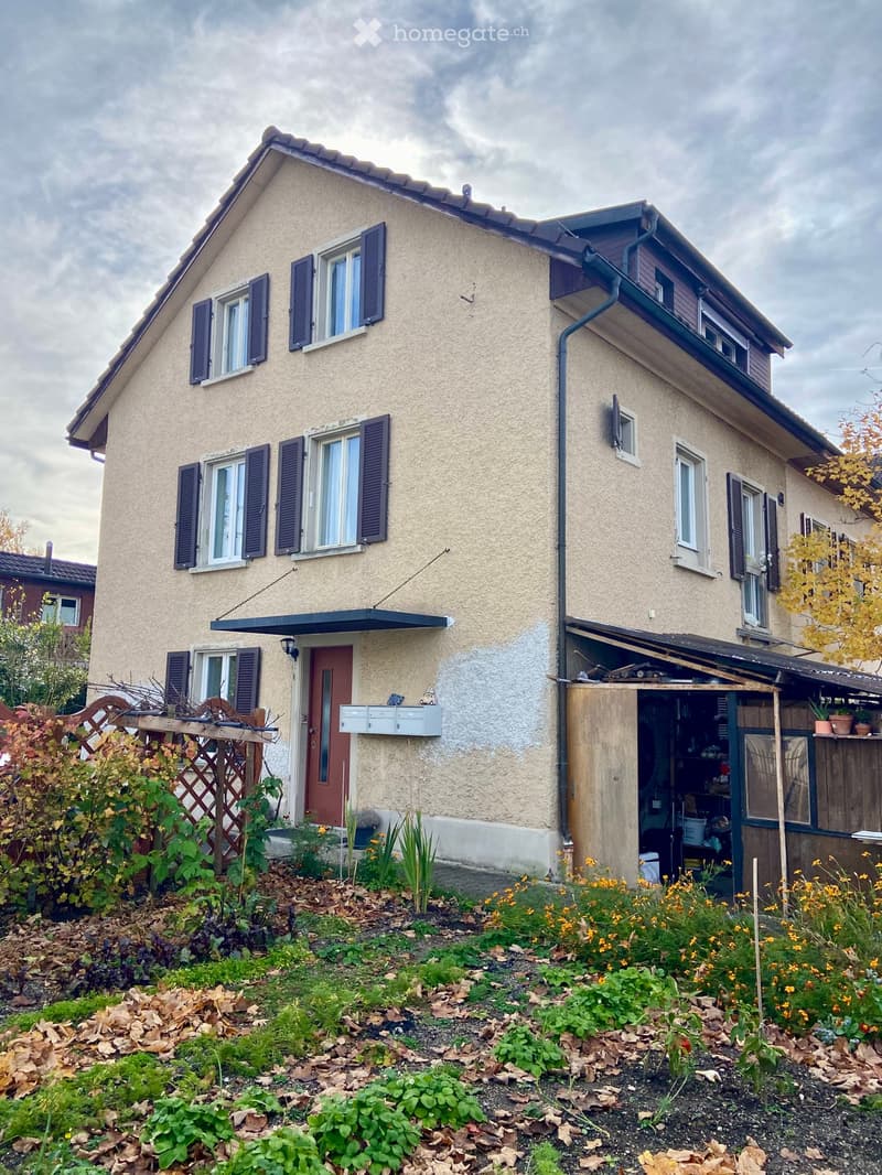 Two-story house with beige siding, multiple windows with shutters, a small garden with flowers and plants, and a covered garage entrance.