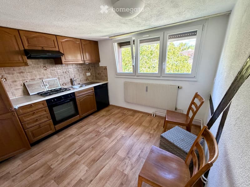 A small kitchen with wooden cabinets, a gas stove, sink, and a few wooden chairs and a stool near the window.