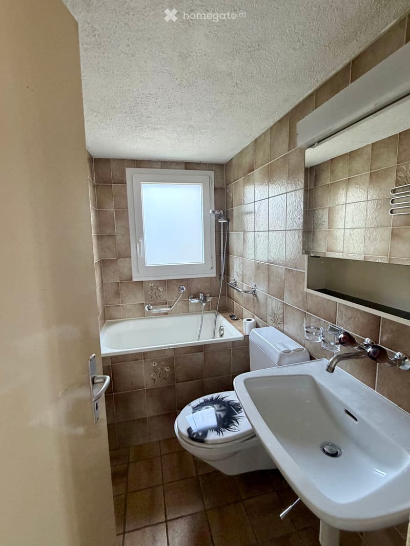 Bathroom with beige tiled walls and floor, featuring a bathtub, toilet, sink, and a window with frosted glass.