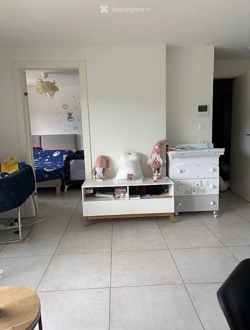 A furnished room with a tiled floor, a white cabinet with toys, a small table with books and toys, a mirror, and a bed visible in the background.