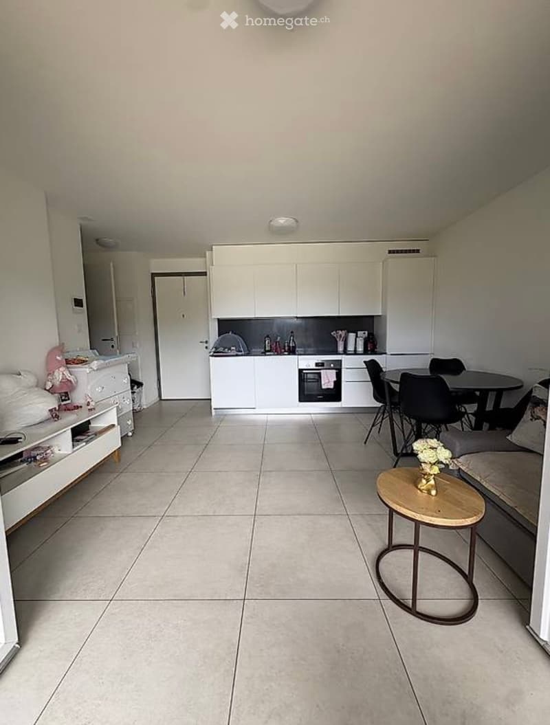 A modern, open-plan living and dining area with a kitchen. The space features a sofa, a small round table with a vase of flowers, a dining table with chairs, and a kitchen area with white cabinets and an oven.