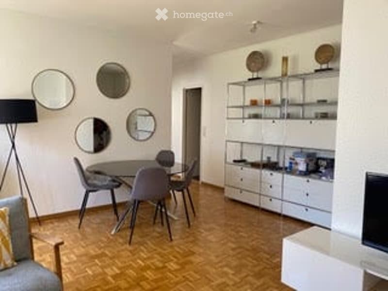 Modern, open-plan living and dining area with light-colored furniture, a grey sofa, a dining table with chairs, a floor lamp, decorative mirrors, and a kitchen area with cabinets and a television.