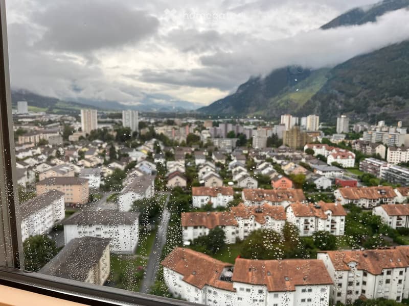 Rainy day view from a balcony or terrace overlooking a town with residential buildings and distant mountains.