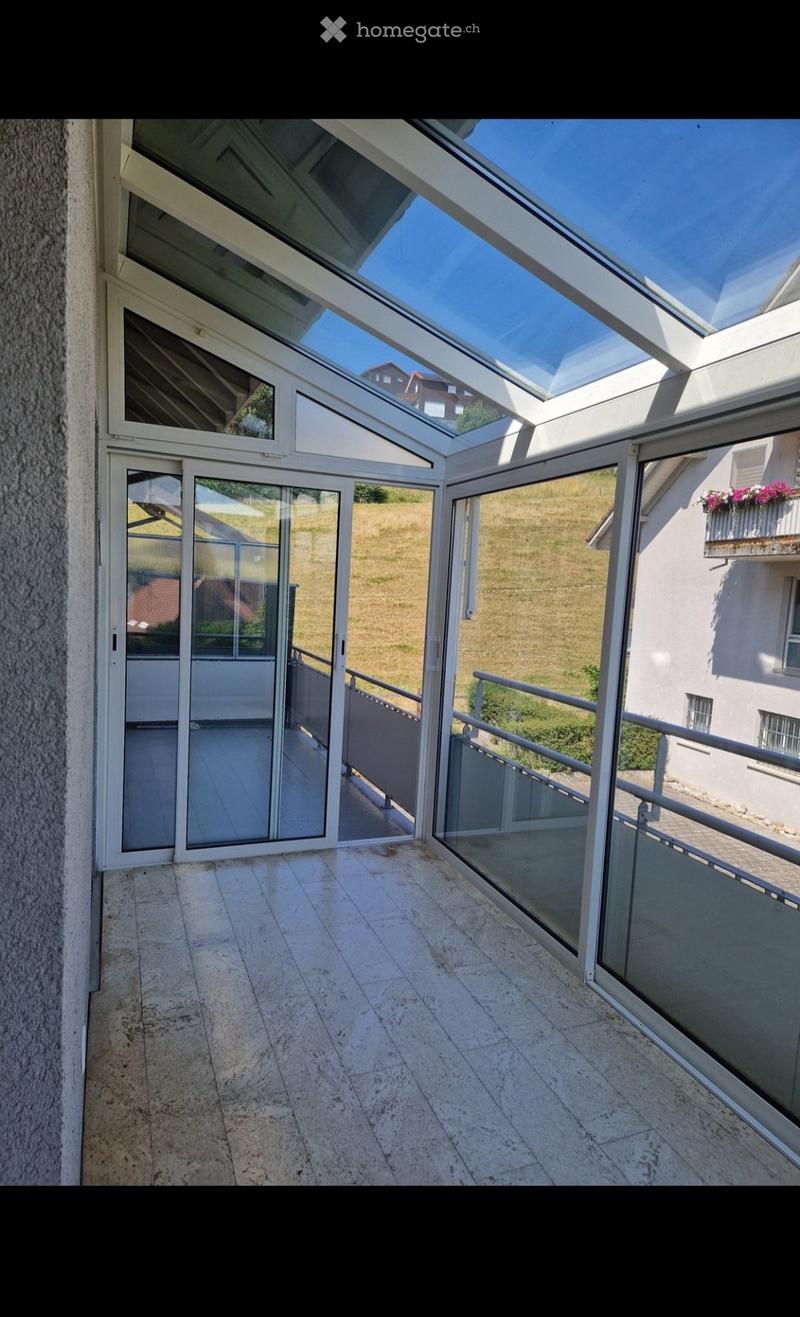 Spacious balcony with a glass roof and sliding glass doors, tiled floor, metal railings, and a view of a grassy area and neighboring building.