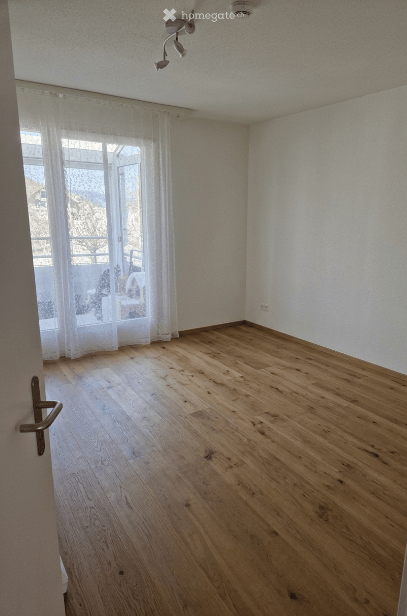 Empty room with wooden flooring, white walls, large window with sheer curtains, and a door handle visible.