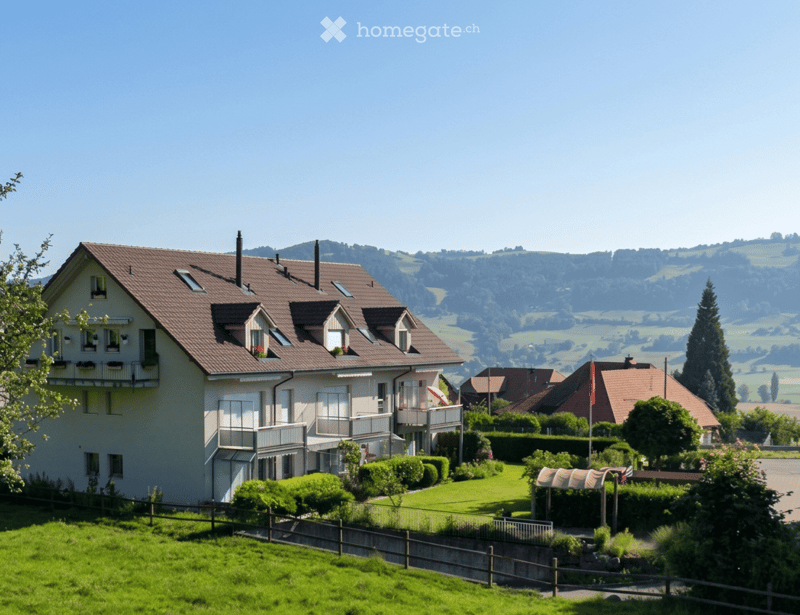 A two-story house with a brown tiled roof, white walls, multiple windows, and balconies. The house is surrounded by a well-maintained lawn, hedges, and trees, with a scenic countryside view in the background.