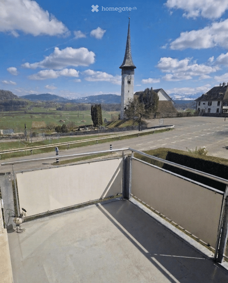 Balcony with metal railing, overlooking a scenic rural landscape with a church tower, fields, and mountains in the background.