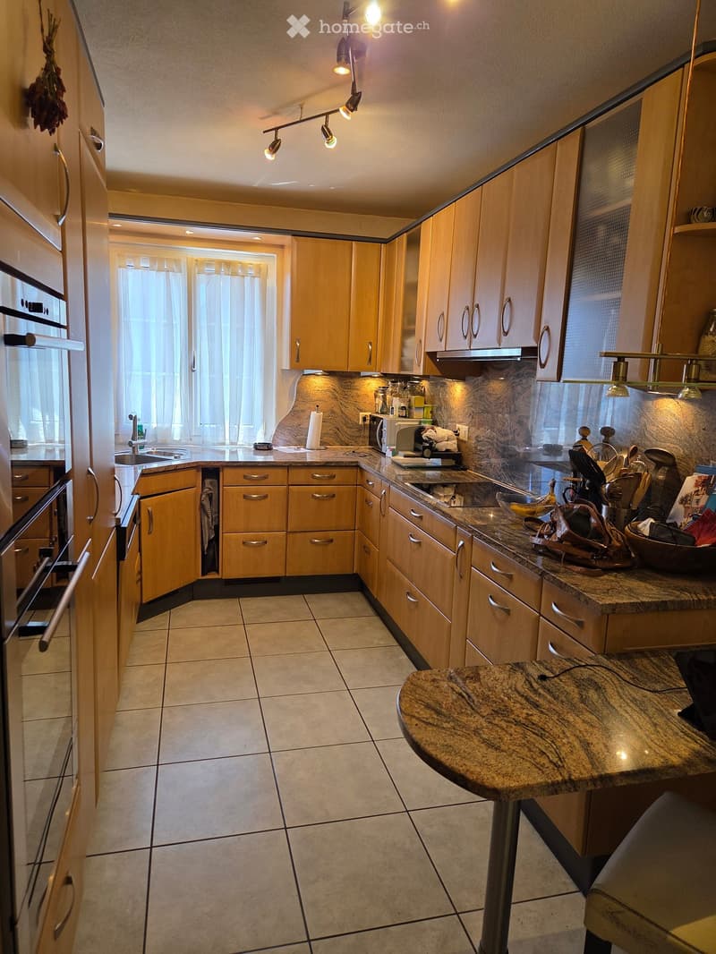 Modern kitchen with light wooden cabinets, granite countertop, stainless steel appliances, and a central island. The space features tiled flooring and ample natural light from a window with curtains.