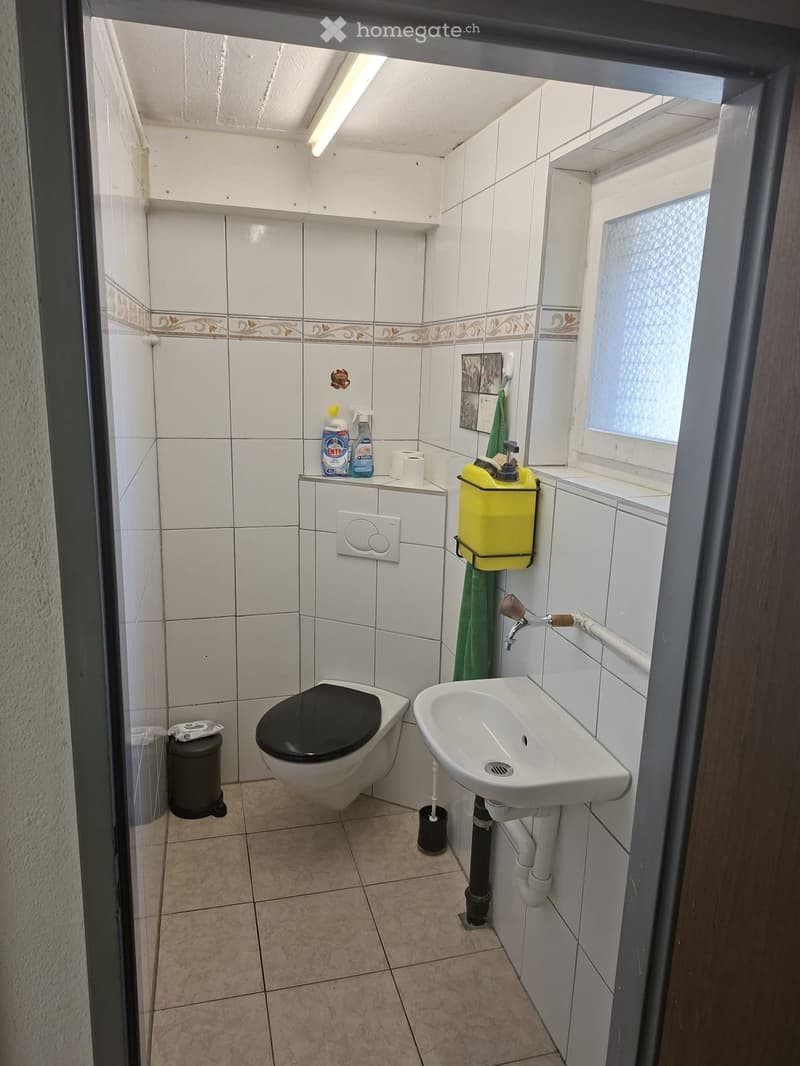 Small bathroom with white tiled walls, a toilet, a sink, a mirror, cleaning supplies, and a towel. The floor is tiled in a light brown color.