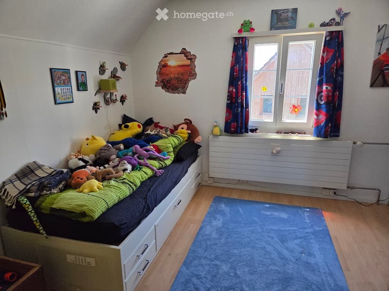 A cozy children's bedroom with a lofted twin-sized bed, colorful stuffed toys, a blue rug, and a window with curtains. The walls feature a unique3D mural and shelves with decorative items.