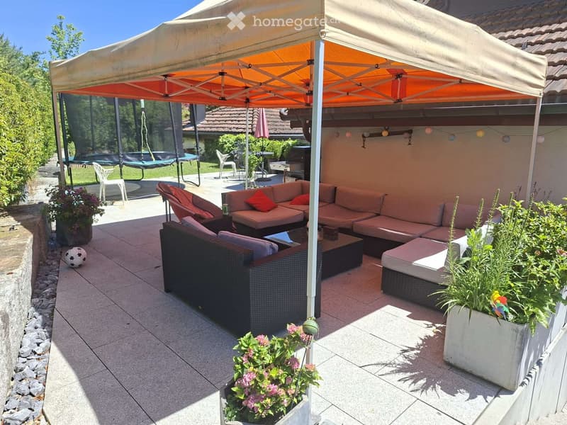 A furnished outdoor patio with a large umbrella, seating area including a sofa, coffee table, and chairs, surrounded by potted plants and a trampoline in the background.