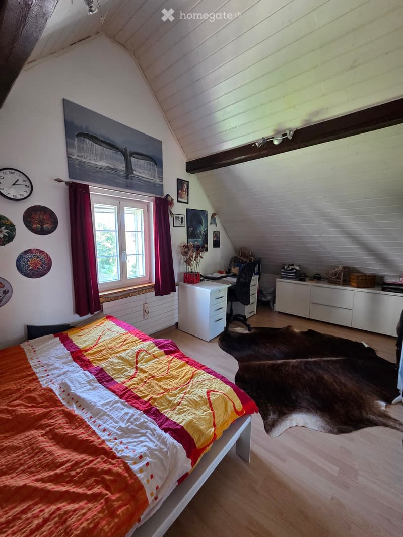 Cozy attic bedroom with slanted wooden ceiling, large window with red curtains, bed with colorful quilt, desk with chair, and built-in white cabinets.