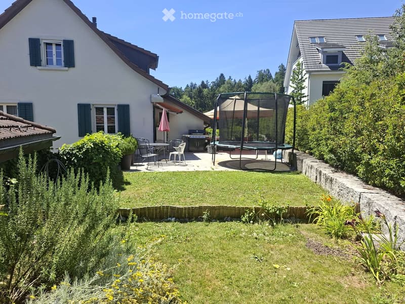 Well-maintained garden with a grassy lawn, trimmed hedges, and a patio area featuring a dining set, umbrella, and trampoline.