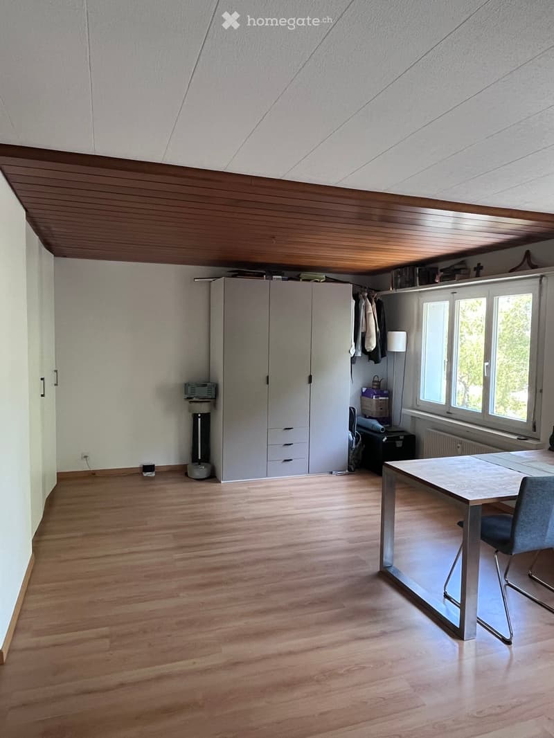 Bright, modern room with wooden flooring, large windows, a white wardrobe, a small table with a chair, and a lamp near the window.