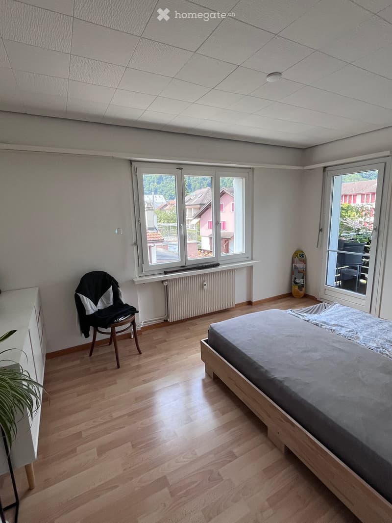 Bright bedroom with wooden flooring, large windows offering a view of the neighborhood, a bed with grey bedding, a wooden chair with a jacket draped over it, a skateboard leaning against the wall, and a small plant near the window.