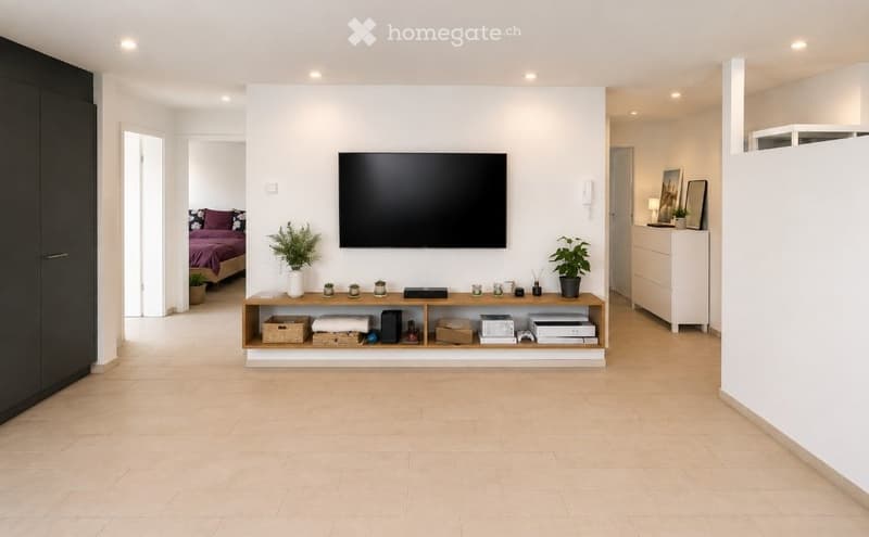 Modern living room with a large flat-screen TV mounted on the wall, a wooden shelf with decorative items, plants, and a small table with a lamp. The room features light-colored tiles and white walls.