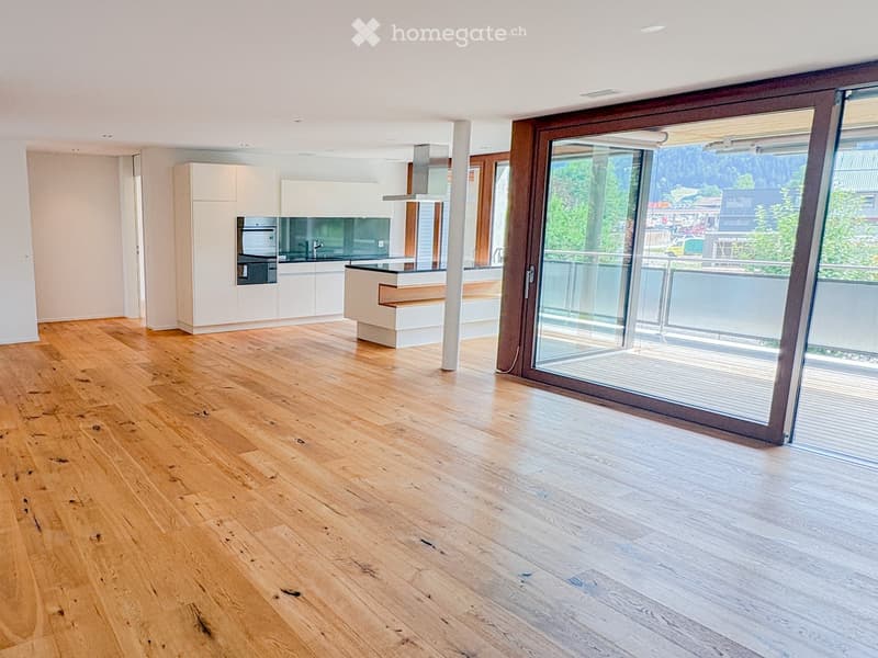 Open-plan space with light wooden flooring, white kitchen cabinets, and a large sliding glass door leading to a balcony with a scenic view.