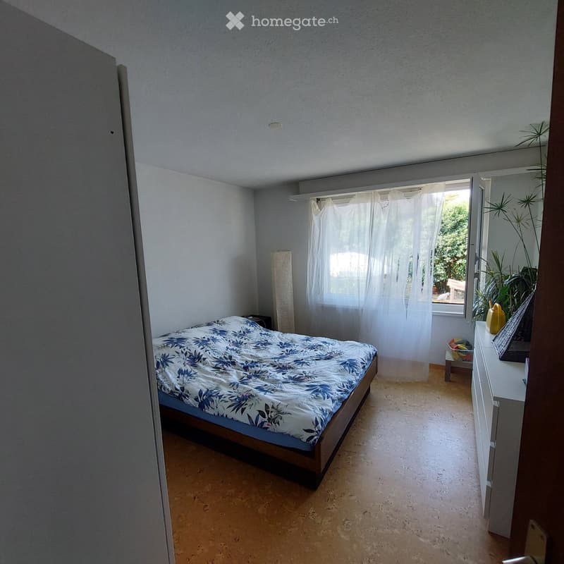 Bedroom with a bed, nightstand, mirror, and a plant near the window. The room has light-colored walls and a wooden floor.