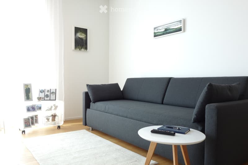 A modern living room with a grey sofa, a white coffee table, a remote control, a book, and picture frames on the wall.