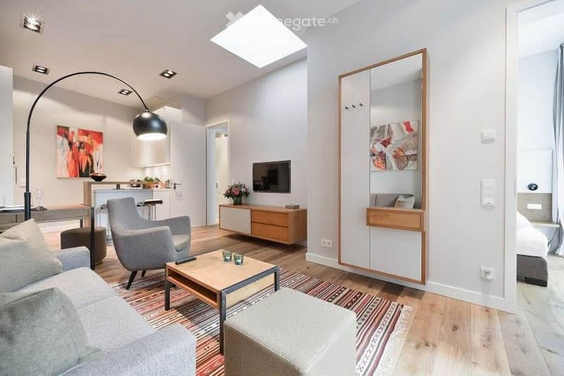 Modern, open-concept living space with a grey sofa, armchair, coffee table, ottoman, floor lamp, large mirror, television, bar counter, and decorative art pieces.