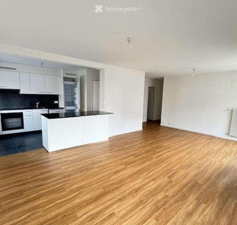 Empty room with wooden flooring, white walls, and a white kitchen area with cabinets and a countertop.