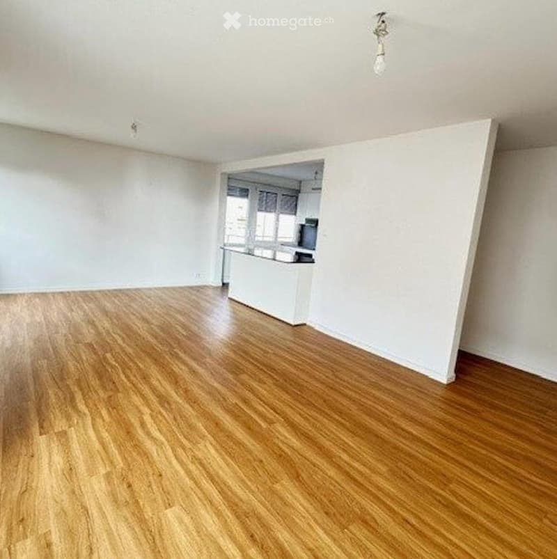 Empty room with wooden flooring and white walls, featuring an open kitchen area in the background.