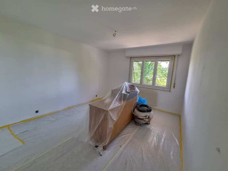 Empty room with white walls, a window, and a radiator. The floor is covered with protective plastic sheeting, and there are some construction materials and tools in the corner.