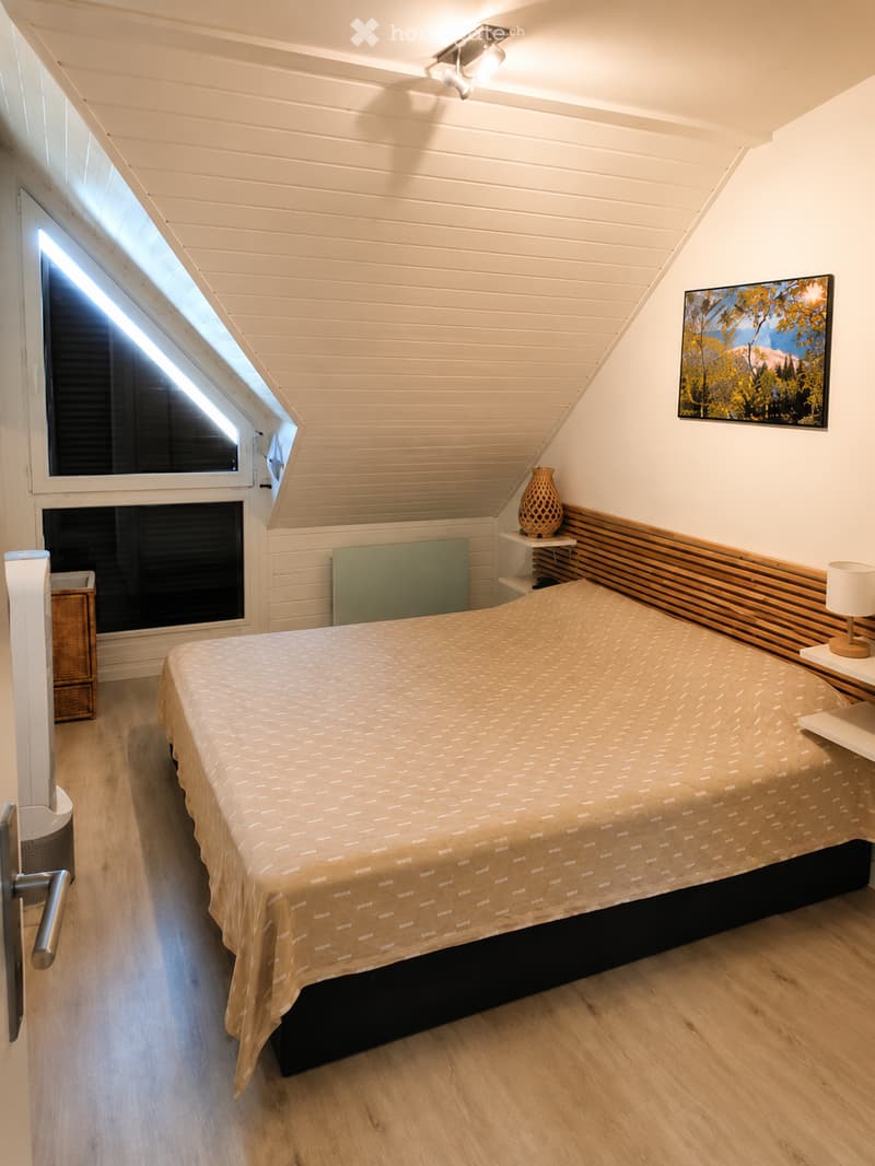 Cozy attic bedroom with wooden slat ceiling, skylight window, bed with patterned duvet, nightstand with lamp, decorative vase, and artwork on wall.