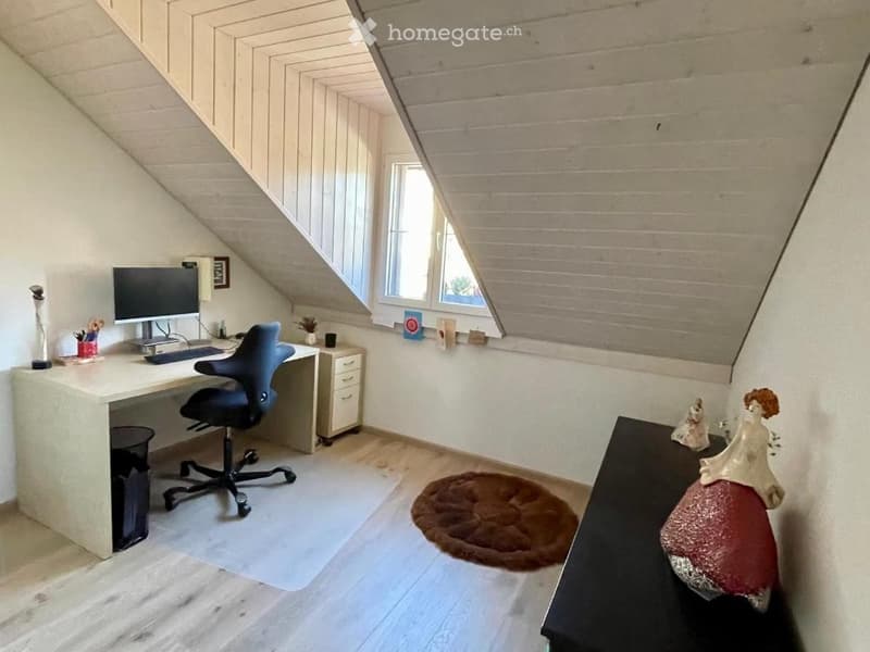 A cozy attic room with wooden walls and slanted ceilings, featuring a desk with a computer, a comfortable chair, a small cabinet, and a decorative table with figurines.